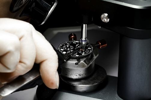 A close-up image of a person working on a watch mechanism using precision tools. The watch face is secured in a metal fixture, and the individual is using a screwdriver to adjust or repair components, indicating a process of intricate craftsmanship.