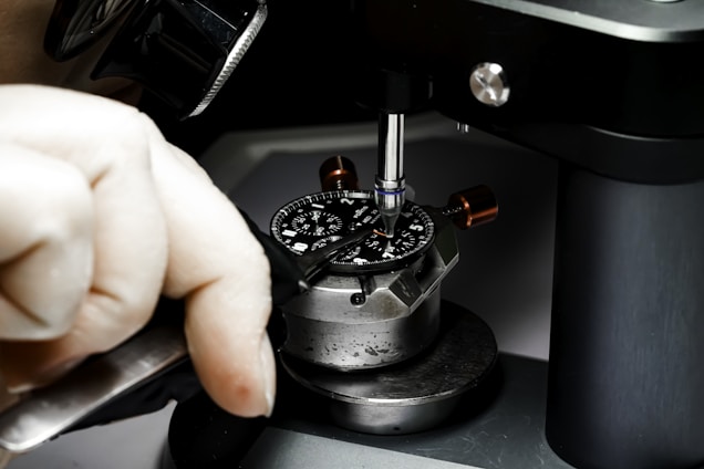 A close-up image of a person working on a watch mechanism using precision tools. The watch face is secured in a metal fixture, and the individual is using a screwdriver to adjust or repair components, indicating a process of intricate craftsmanship.