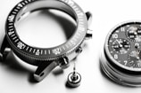 A detailed close-up of disassembled watch parts, including a metallic bezel with marked increments, a round watch face with multiple chronograph dials and visible numerals, and a small screw crown. The components are intricately arranged on a smooth, white surface, highlighting their precision engineering.