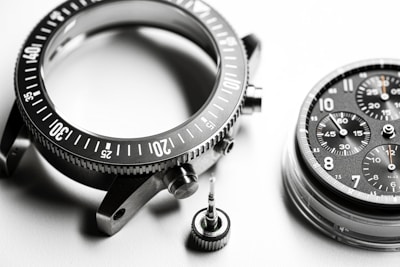 A detailed close-up of disassembled watch parts, including a metallic bezel with marked increments, a round watch face with multiple chronograph dials and visible numerals, and a small screw crown. The components are intricately arranged on a smooth, white surface, highlighting their precision engineering.