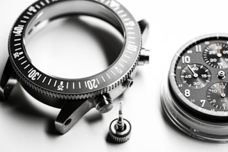 A detailed close-up of disassembled watch parts, including a metallic bezel with marked increments, a round watch face with multiple chronograph dials and visible numerals, and a small screw crown. The components are intricately arranged on a smooth, white surface, highlighting their precision engineering.