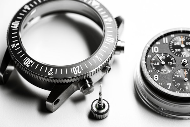 A detailed close-up of disassembled watch parts, including a metallic bezel with marked increments, a round watch face with multiple chronograph dials and visible numerals, and a small screw crown. The components are intricately arranged on a smooth, white surface, highlighting their precision engineering.