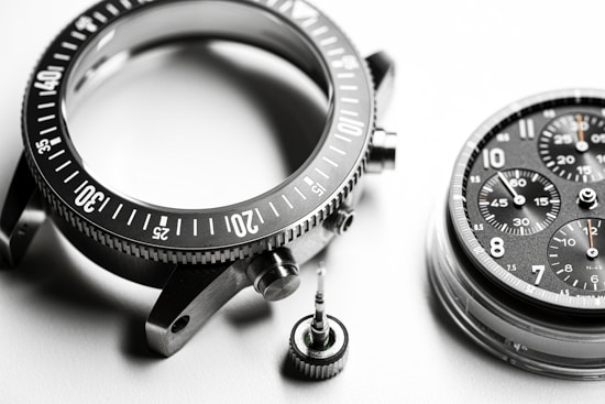 A detailed close-up of disassembled watch parts, including a metallic bezel with marked increments, a round watch face with multiple chronograph dials and visible numerals, and a small screw crown. The components are intricately arranged on a smooth, white surface, highlighting their precision engineering.