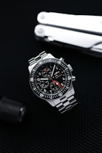 A sleek stainless steel wristwatch with a black dial and three subdials is positioned against a dark textured background. The watch features white numerals and markings with a red-tipped second hand. A multitool, partially visible, is placed in the background, adding a rugged, utilitarian element.