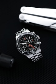 A sleek stainless steel wristwatch with a black dial and three subdials is positioned against a dark textured background. The watch features white numerals and markings with a red-tipped second hand. A multitool, partially visible, is placed in the background, adding a rugged, utilitarian element.