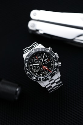 A sleek stainless steel wristwatch with a black dial and three subdials is positioned against a dark textured background. The watch features white numerals and markings with a red-tipped second hand. A multitool, partially visible, is placed in the background, adding a rugged, utilitarian element.