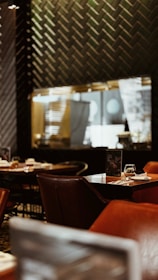 A cozy restaurant interior with dim lighting. The tables are set with empty wine glasses and leather chairs. The wall has a herringbone pattern, and there are reflections visible in the mirror, contributing to a warm and intimate atmosphere.