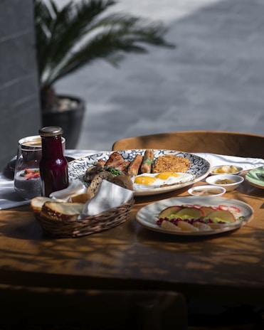 A table set with a variety of breakfast foods, including fried eggs, sausages, grilled meats, and bread in a basket. There are accompaniments like sliced fruits, small bowls of dips or spreads, and a glass of chia pudding with strawberries. A dark bottle, possibly juice or beverage, rests near the plate. The setting suggests an outdoor or brightly lit indoor dining area with a blurred plant in the background.