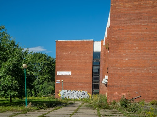 A large brick building with graffiti on its lower section is surrounded by greenery and a clear blue sky. A sign with Cyrillic text is visible on the building. There is a path leading to the building, lined with spherical lamps on poles. Vegetation appears overgrown near the pathway.