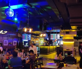 A lively indoor bar setting with a dimly lit atmosphere and vibrant neon lighting. Several people are seated at tables socializing and enjoying drinks. A disco ball hangs from the ceiling, and screens displaying various menus are visible above the bar. The bar counter is illuminated, and the interior decor features wood and industrial elements.