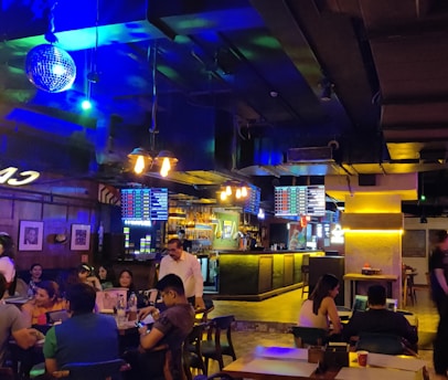A lively indoor bar setting with a dimly lit atmosphere and vibrant neon lighting. Several people are seated at tables socializing and enjoying drinks. A disco ball hangs from the ceiling, and screens displaying various menus are visible above the bar. The bar counter is illuminated, and the interior decor features wood and industrial elements.