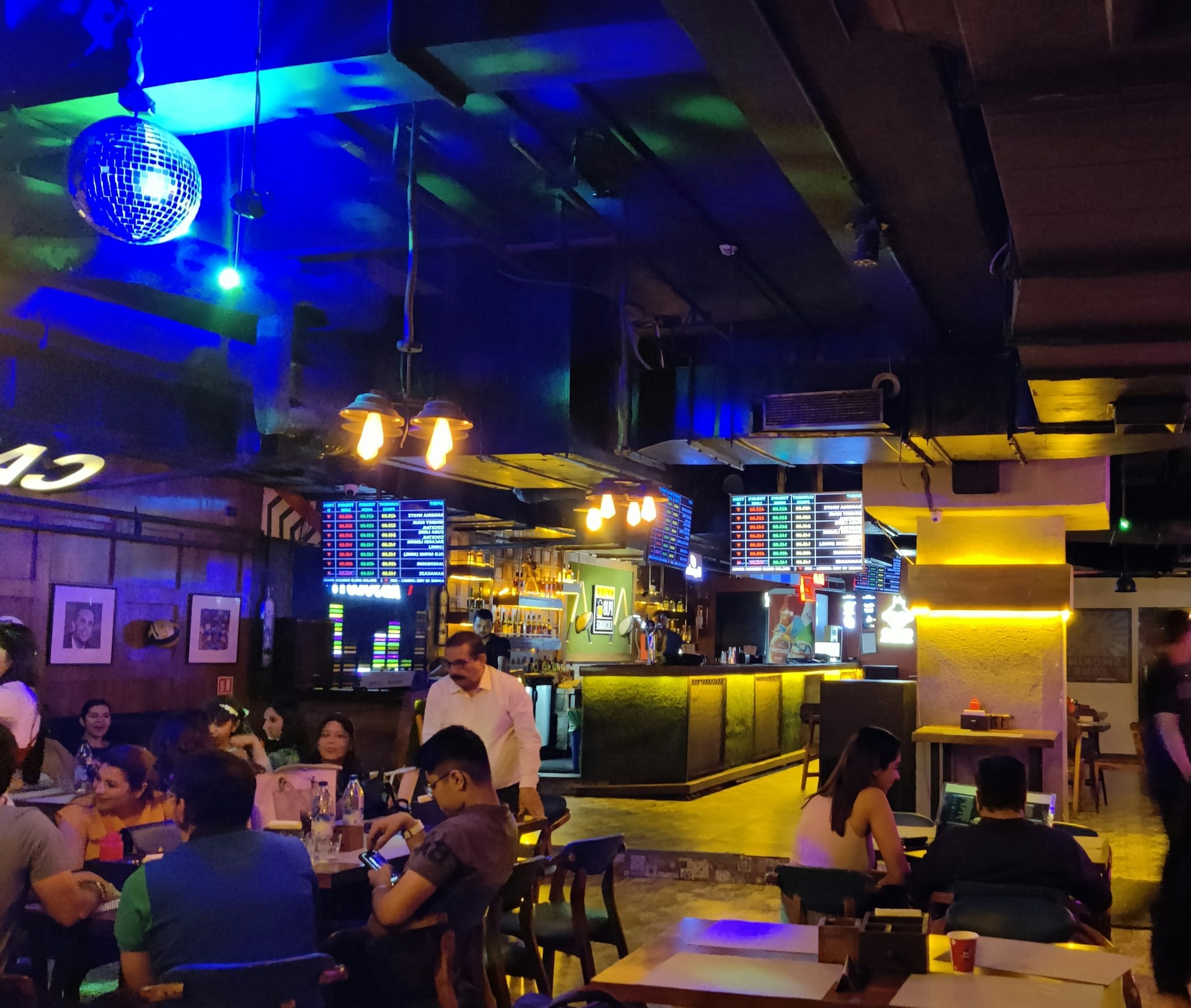 A lively indoor bar setting with a dimly lit atmosphere and vibrant neon lighting. Several people are seated at tables socializing and enjoying drinks. A disco ball hangs from the ceiling, and screens displaying various menus are visible above the bar. The bar counter is illuminated, and the interior decor features wood and industrial elements.