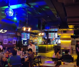 A lively indoor bar setting with a dimly lit atmosphere and vibrant neon lighting. Several people are seated at tables socializing and enjoying drinks. A disco ball hangs from the ceiling, and screens displaying various menus are visible above the bar. The bar counter is illuminated, and the interior decor features wood and industrial elements.