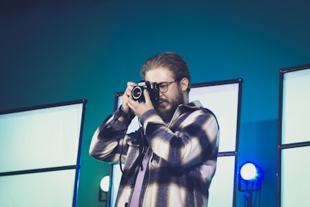 A person is taking a photograph with a camera while standing in front of a backdrop illuminated by bright lights. The individual is wearing glasses and a plaid jacket, appearing focused on capturing the shot.