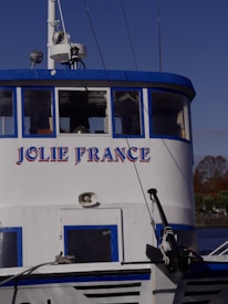 The image features the front section of a white and blue boat with the name 'Jolie France' written on it. The boat has several windows and a mast with equipment on top. It's docked near a body of water with trees and infrastructure visible in the background under a clear blue sky.