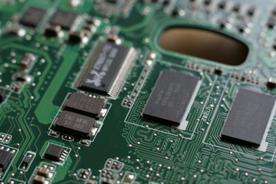 A close-up view of a green circuit board featuring various components including integrated circuits, resistors, and capacitors interconnected with intricate pathways. The surface is covered with small, rectangular black chips marked with white text, and metallic connections. The focus highlights the complex design typical of an electronic device's internal structure.