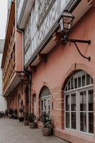A narrow alleyway with traditional German half-timbered architecture. The buildings have a mix of pink and beige facades with large arched windows. Black metal lanterns are mounted on the walls, and potted plants in wicker baskets line the side of the building along the cobblestone path.