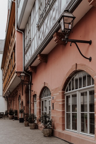 A narrow alleyway with traditional German half-timbered architecture. The buildings have a mix of pink and beige facades with large arched windows. Black metal lanterns are mounted on the walls, and potted plants in wicker baskets line the side of the building along the cobblestone path.