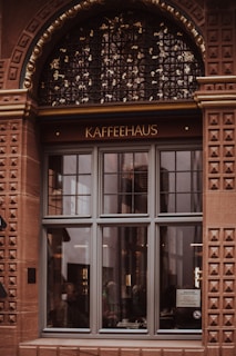 A detailed facade of a building with a large arched window displaying the word 'KAFFEEHAUS'. The window is framed in intricate brickwork with a decorative black grille above it. People can be seen through the window sitting inside, with reflections on the glass.