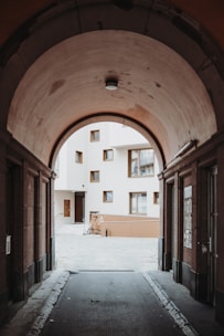 A vaulted walkway leads to an open courtyard surrounded by modern white and beige buildings with several windows. The archway creates a frame around the view of the courtyard, adding depth to the architectural setting.