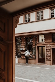 A person stands at a street-side order window. The scene includes festive holiday decorations, with garlands around the window. The building has a rustic brick facade and several windows, set within a paved narrow urban setting.