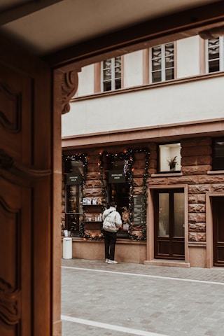 A person stands at a street-side order window. The scene includes festive holiday decorations, with garlands around the window. The building has a rustic brick facade and several windows, set within a paved narrow urban setting.