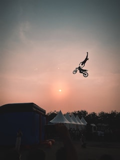 A motorcyclist is performing a mid-air stunt against the backdrop of a dusky sky with the sun partially visible. The rider is suspended in the air with their silhouette sharply contrasted by the warm tones of the sunset. Below, there are tents and a small crowd of people gathered, possibly at an outdoor event.