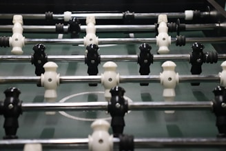A foosball table with rows of molded players attached to rods, featuring alternating black and white figures. The field is green with marked white lines and goals at each end.