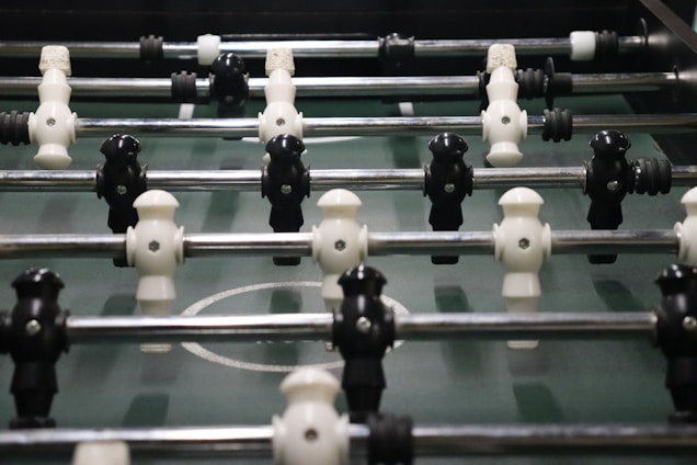 A foosball table with rows of molded players attached to rods, featuring alternating black and white figures. The field is green with marked white lines and goals at each end.