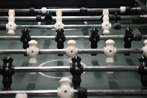 A foosball table with rows of molded players attached to rods, featuring alternating black and white figures. The field is green with marked white lines and goals at each end.