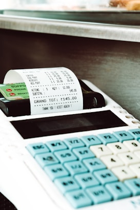 A close-up of a payment terminal with a printed receipt. The receipt includes various purchased items such as coffee, egg puff, and biscuits, with individual prices listed. The total amount of 145.00 is shown. The keypad of the terminal with blue and white keys is visible.