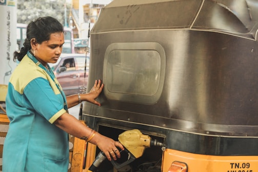 A woman in a uniform is refueling an auto-rickshaw at a gas station. She is holding a fuel nozzle, and there is another vehicle visible in the background through the window.