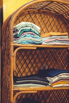 Tightly stacked folded clothes in various colors are neatly arranged on a two-tiered rattan shelf. The bamboo structure features a curved top and lattice design on the sides and back, set against a yellow wall.
