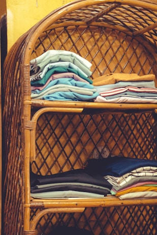 Tightly stacked folded clothes in various colors are neatly arranged on a two-tiered rattan shelf. The bamboo structure features a curved top and lattice design on the sides and back, set against a yellow wall.