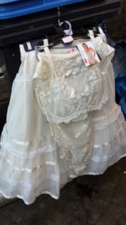 A white lace wedding or bridal dress is hanging on a black hanger in front of a blue trash bin. The dress includes intricate lace details and layered fabric. A tag with an image of a model wearing a similar garment is attached to the hanger.