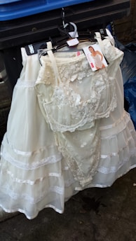 A white lace wedding or bridal dress is hanging on a black hanger in front of a blue trash bin. The dress includes intricate lace details and layered fabric. A tag with an image of a model wearing a similar garment is attached to the hanger.