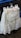 A white lace wedding or bridal dress is hanging on a black hanger in front of a blue trash bin. The dress includes intricate lace details and layered fabric. A tag with an image of a model wearing a similar garment is attached to the hanger.