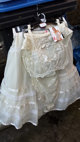 A white lace wedding or bridal dress is hanging on a black hanger in front of a blue trash bin. The dress includes intricate lace details and layered fabric. A tag with an image of a model wearing a similar garment is attached to the hanger.
