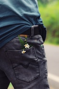 A person is wearing distressed denim jeans with small white flowers and green leaves tucked inside the back pocket. The jeans contrast with a blue shirt. The background suggests an outdoor setting with greenery visible.