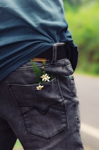 A person is wearing distressed denim jeans with small white flowers and green leaves tucked inside the back pocket. The jeans contrast with a blue shirt. The background suggests an outdoor setting with greenery visible.