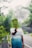 A person wearing a California-themed t-shirt is standing on a road surrounded by lush green foliage. They are holding up a large fern leaf in front of their face. The background features a misty atmosphere with more trees and parts of the road visible.