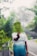 A person wearing a California-themed t-shirt is standing on a road surrounded by lush green foliage. They are holding up a large fern leaf in front of their face. The background features a misty atmosphere with more trees and parts of the road visible.