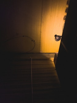 A dimly lit room features a single light source casting a warm, yellow glow on the wooden wall. A dark cable is draped loosely against the wall, and a standing lamp is partially visible. The light creates long shadows, enhancing the atmosphere.