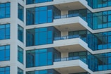 The image features a modern high-rise apartment building with several floors. The facade is characterized by large glass windows and beige walls. Each floor has a balcony protected by dark railings, and the architectural lines create a repeating pattern.