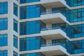 The image features a modern high-rise apartment building with several floors. The facade is characterized by large glass windows and beige walls. Each floor has a balcony protected by dark railings, and the architectural lines create a repeating pattern.