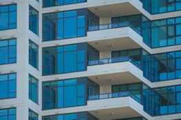 The image features a modern high-rise apartment building with several floors. The facade is characterized by large glass windows and beige walls. Each floor has a balcony protected by dark railings, and the architectural lines create a repeating pattern.