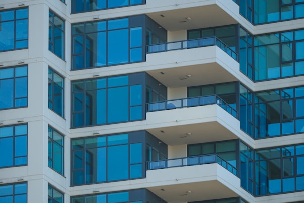 The image features a modern high-rise apartment building with several floors. The facade is characterized by large glass windows and beige walls. Each floor has a balcony protected by dark railings, and the architectural lines create a repeating pattern.