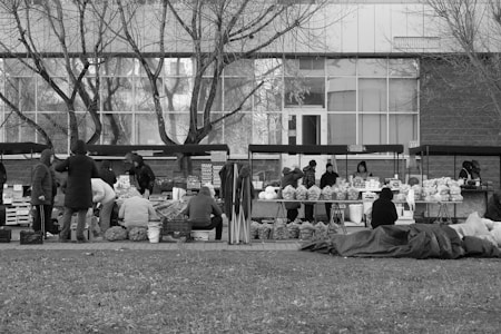 A black and white outdoor market scene with several vendors and shoppers. The vendors have set up tables with various goods including fruits and vegetables. People are dressed in warm clothing, suggesting cold weather. Trees with bare branches stand in the background, reflecting a winter setting.