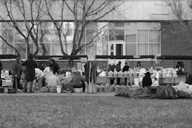 A black and white outdoor market scene with several vendors and shoppers. The vendors have set up tables with various goods including fruits and vegetables. People are dressed in warm clothing, suggesting cold weather. Trees with bare branches stand in the background, reflecting a winter setting.