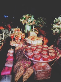 A beautifully decorated dessert table featuring a variety of treats, including cupcakes with cream tops placed on a stand, wrapped bags of popcorn, and multiple decorative gift boxes. Several glasses of a vibrant pink beverage are also visible, along with lush floral arrangements in vases enhancing the rich, celebratory atmosphere.